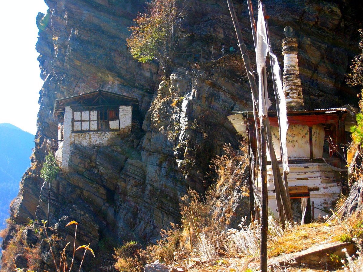 A small stone hut and whitewashed memorial stupa on a steep forested mountainside in Bhutan, hung with prayer flags.