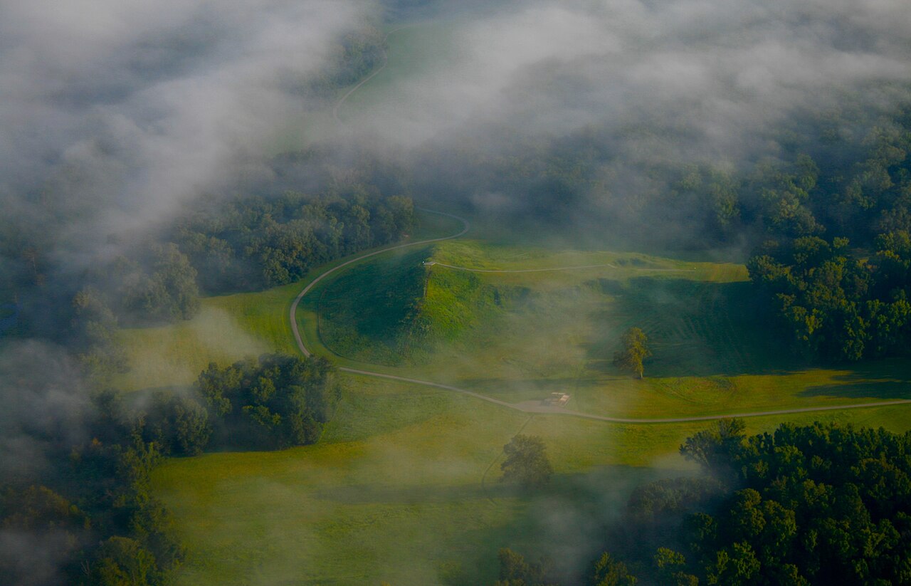 Aerial view through low mist of the earthworks at Poverty Point, Louisiana, showing a large grassed mound and curving ridges.